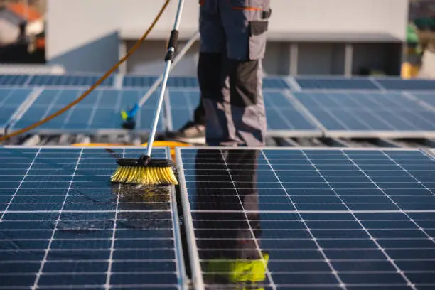 Un technicien nettoie des panneaux solaires installés sur un toit à l'aide d'une brosse télescopique jaune. De la mousse d'eau est visible sur les panneaux nettoyés.