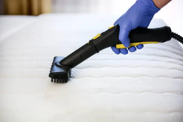 Une main gantée de bleu utilise une buse de nettoyage à vapeur noire et jaune pour nettoyer en profondeur un matelas blanc matelassé.