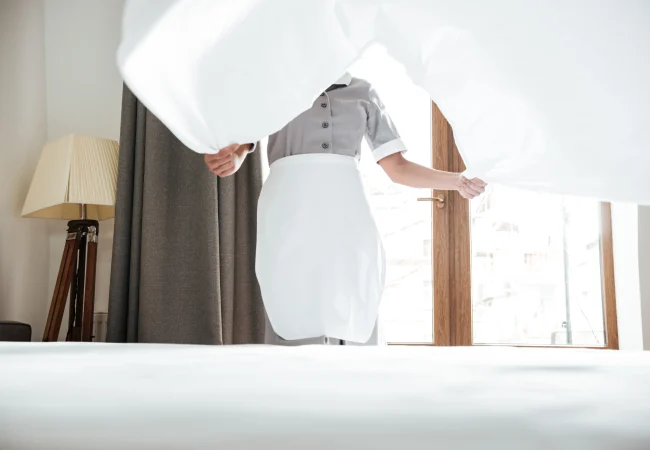 Une femme de chambre en uniforme gris et tablier blanc secouant un drap blanc pour faire un lit d'hôtel près d'une fenêtre lumineuse.
